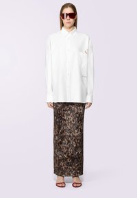 Camicia bianca oversize con bottoni e tasca sul petto, abbinata a una maxi gonna leopardata marrone. Gli occhiali da sole rossi e sandali semplici completano il look.