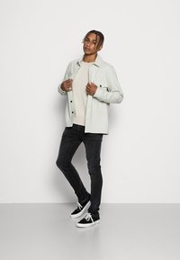 Casaco cinza claro, abotoado, sobre um suéter bege, combinado com jeans pretos e sapatilhas pretas. Tecido de acabamento natural com dois bolsos frontais.