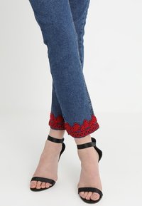 Blå denimjeans med röda broderade fålldetaljer, tillsammans med svarta remsandaler, som visar en jämn textur och en figurnära design.