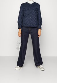 Giacca navale trapuntata con due tasche sul petto, colletto tondo e polsini elasticizzati, abbinata a jeans scuri larghi e sneakers bianche.