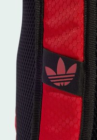 Czerwona i czarna plecak z heksagonalnym teksturowanym materiałem, czarnym paskiem z wytłoczonym logo w postaci trzech pasków oraz akcentami z siateczki.