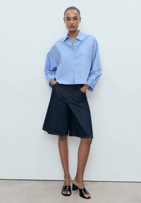 Lichtblauw, collared button-up shirt met oversized mouwen, gecombineerd met donkere denim kniebroekshorts en zwarte open-toe schoeisel.