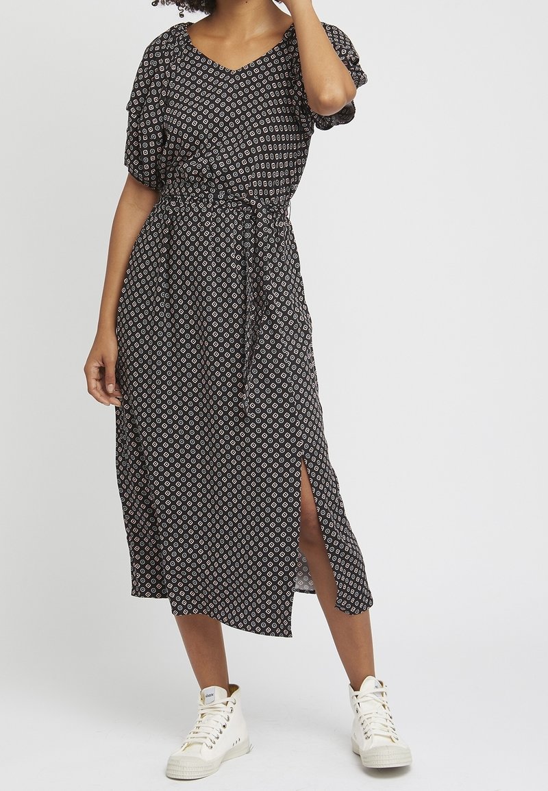 Zwarte jurk met patroon, een V-hals, korte mouwen en een elastische taille. Heeft een zijspleet en wordt gecombineerd met witte sneakers.