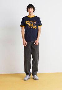 Marineblaues T-Shirt mit gelben Akzenten und grafischem Druck, kombiniert mit dunkelgrauen Hosen und grauen Turnschuhen. Einfaches, lässiges Design.
