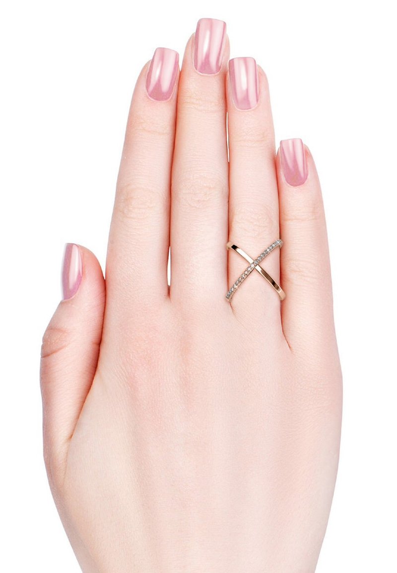 Anello incrociato in oro con una piccola fila di pietre trasparenti su un lato; indossato su una mano con unghie lucide rosa chiaro su uno sfondo bianco.