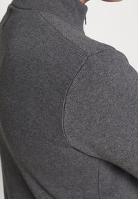 Detailní záběr na osobu, která má na sobě tmavě šedý pletený svetr s vysokým límcem a viditelnými švy na rameni.