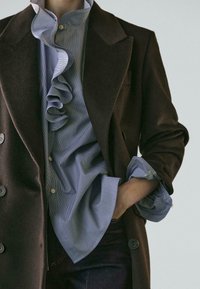 Osoba nosící hnědý dvouřadý kabát přes modrou halenku s volánky a vyhrnutými rukávy, s jednou rukou zasunutou do kapsy tmavých kalhot.