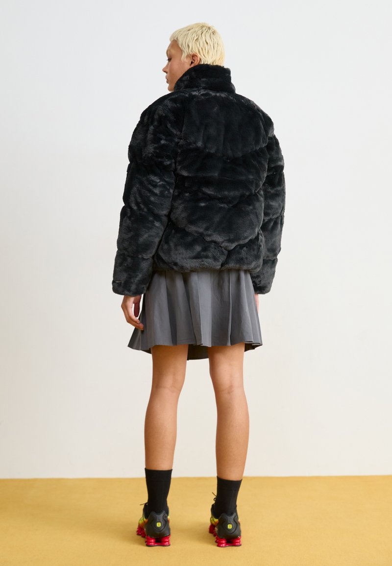 Doudoune en fausse fourrure noire avec un col haut et un design matelassé, associée à une jupe plissée grise et des chaussures de sport noires.