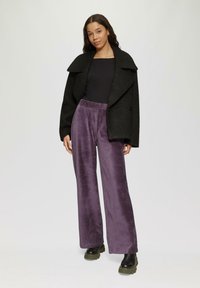 Zwarte wolblend jas met brede revers, gedragen over een zwarte top. Paarse fluwelen wijde broek en zwarte platformlaarzen maken de outfit compleet.
