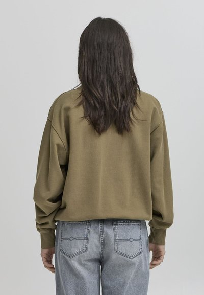 Maglione oversized verde oliva con spalle cadenti, polsini a costine e texture morbida. Abbinato a jeans azzurri chiari strappati con dettagli sulle tasche posteriori.