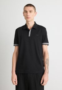 Polo preto com colarinho e detalhes das mangas em cinza. Apresenta um design minimalista e o logótipo 'BOSS' no lado esquerdo. Textura suave.