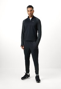 Schwarzes sportliches Viertel-Reißverschluss-Oberteil mit strukturierten Ärmeln, kombiniert mit schwarzen Jogginghosen, die eine Tasche und gemusterte Unterschenkel aufweisen. Schwarze Sneakers.