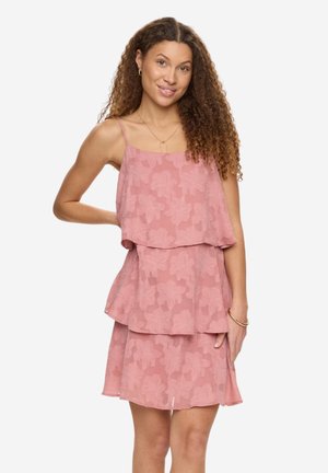 Jeune femme aux cheveux bouclés portant une robe rose à volants sans manches avec des motifs floraux, souriante et posant avec une main sur la hanche.
