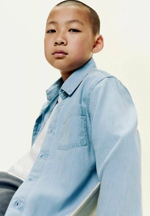 Chemise en denim bleu clair avec col, grande poche avant et fermeture par boutons, portée sur une chemise blanche, présentée sur un fond neutre.