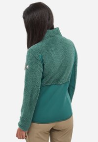 Frau mit langen dunklen Haaren, die eine türkisfarbene Fleecejacke mit strukturiertem oberen Rücken und Ärmeln trägt, steht vor einem weißen Hintergrund.