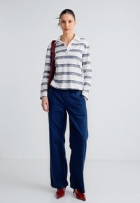 Polo de manga larga a rayas en blanco con franjas azules y moradas, combinado con pantalones anchos azules y zapatos de tacón altos rojos.