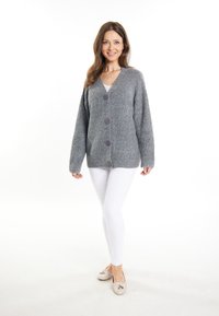 Cardigan grigio in lana con scollo a V, caratterizzato da tre bottoni viola e lavorazione a maglia texture. Indossato sopra una maglietta bianca e abbinato a pantaloni bianchi.