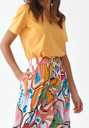 Kobieta ubrana w musztardowożółtą bluzkę z krótkim rękawem i dekoltem w serek oraz spódnicę z wysokim stanem w wielokolorowy abstrakcyjny wzór z troczkiem w pasie.