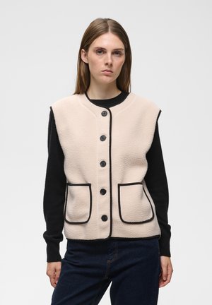 Femme portant un gilet beige boutonné avec des bordures noires sur un haut noir à manches longues et un jean foncé, debout devant un fond uni.