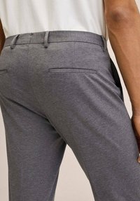 Man in grijze, nauwsluitende pantalon en een wit shirt, zijaanzicht van de taille tot halverwege de dijen, met zakken en riemlussen.