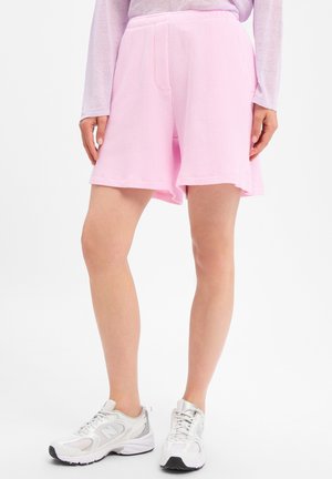 Persoon die lichtroze gestructureerde shorts en witte sportschoenen draagt, staand op een witte achtergrond.