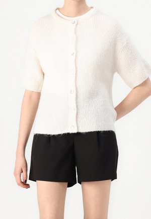 Persoon draagt een wit gebreid vest met korte mouwen en knopen, gecombineerd met losse zwarte shorts, zichtbaar van schouders tot halverwege de dijen.