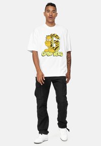 Camiseta de algodón blanca con un gráfico colorido de Garfield que incluye una gran "G" y rayas negras y amarillas. Combinada con pantalones cargo negros y zapatillas blancas.