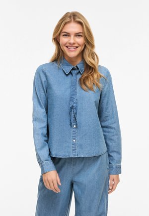 Lächelnde Frau mit langen blonden Haaren, die ein hellblaues Jeanshemd mit kleinem Knoten-Detail und passende Jeanshose trägt.