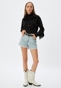 Zwarte suède jas met twee borstzakken en franjes, gecombineerd met lichtblauwe versleten denim shorts versierd met zilveren studs en witte cowboylaarzen.