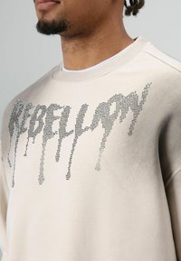 Beige sweatshirt met een geribde kraag, voorzien van rhinestone letters die het woord "REBELLION" spellen in een druppelend ontwerp. Gladde textuur.