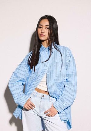 Jeune femme aux longs cheveux foncés portant une chemise oversize bleu clair à rayures, par-dessus un crop top blanc et un jean bleu clair, posant les mains sur les hanches.