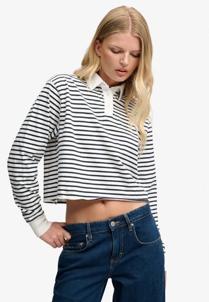 Lyshåret kvinde iført en hvid og sort stribet cropped langærmet skjorte og blå jeans, der kigger ned med hånden på hoften.