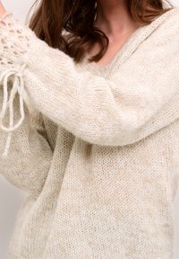 Sweter dziewiarski w jasnym beżu, z luźnym krojem, kapturem i akcentami w postaci sznurka. Teksturalna przędza o miękkim, puszystym wyglądzie.
