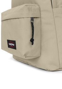 Zaino beige con texture in tessuto. Presenta una tasca frontale con una cerniera e un cordino, oltre a una toppa con logo nera. Design minimalista.