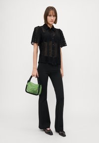 Camicia nera in pizzo con maniche a sbuffo e bottoni, abbinata a pantaloni neri svasati e una borsa di perline verdi con manico nero.