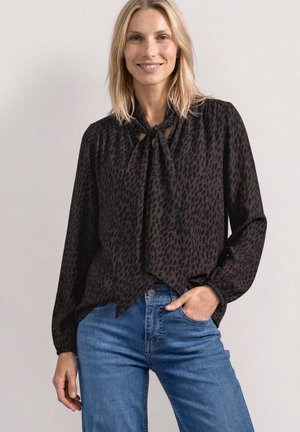 Kvinde med blondt hår iført sort bluse med lange ærmer og sløjfe ved halsen samt blå jeans, smilende med den ene hånd i lommen, lyst baggrund.