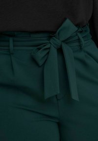 Dark green high-waist pants with pleats and a wide fabric belt tied in a bow at the waist, paired with a black top.