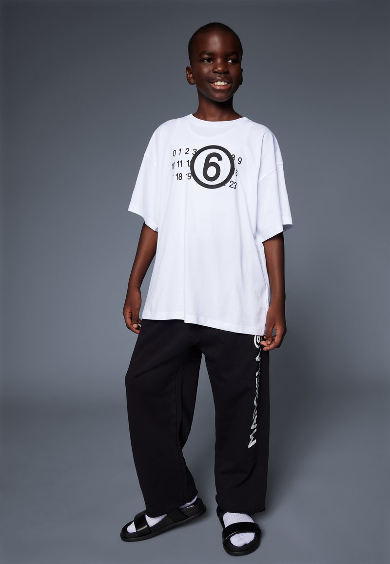 T-shirt bianca oversize con una grafica circolare nera che mostra il numero 6, abbinata a pantaloni della tuta neri con testo logo bianco e sandali.