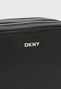 Μαύρη δερμάτινη τσάντα με υφή, με ασημένια φερμουάρ και ασημένιο λογότυπο DKNY. Συμπαγές ορθογώνιο σχήμα με λιτές λεπτομέρειες.