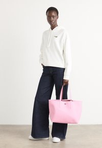 Vit huvtröja, blå vida jeans och en rosa läderhandväska med logotypdetalj. Modell som bär vita sneakers. Minimal bakgrund.
