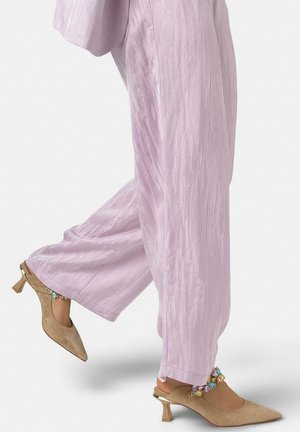 Persona que lleva pantalones anchos texturizados de color púrpura claro y zuecos beige de tacón bajo con punta puntiaguda y tiras para el tobillo con piedras preciosas de colores.