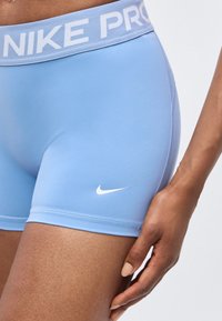 Short Nike Pro bleu clair avec logo blanc sur la cuisse porté par une personne à la peau brune et la main reposant près du short.
