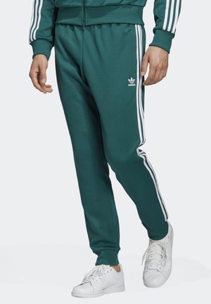 Person trägt grüne Adidas Trainingshose mit weißen Streifen und Logo, kombiniert mit weißen Sneakers, stehend vor einem einfarbigen Hintergrund.