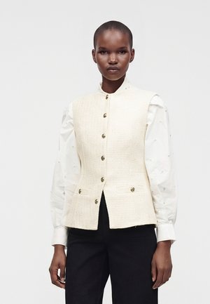 Femme aux cheveux courts porte un gilet texturé crème boutonné sur un chemisier blanc à manches longues avec des détails en perles et un pantalon noir.
