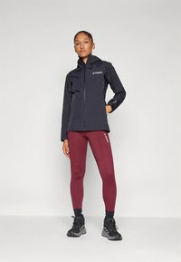 Femme debout portant une veste zippée noire, un legging bordeaux, des chaussettes noires et des chaussures de randonnée noires sur un fond clair uni.