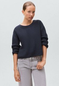 Pulover tricotat de culoare bleumarin închis, cu guler rotund și manșete răsucite, asortat cu jeansi gri deschis. Textura este în relief, cu detalii subtile.