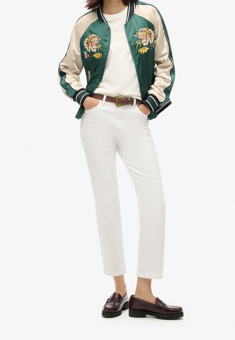 Femme portant une veste bomber en satin vert et crème avec broderie tigre, pantalon blanc, chemise blanche, ceinture marron et mocassins marron foncé.