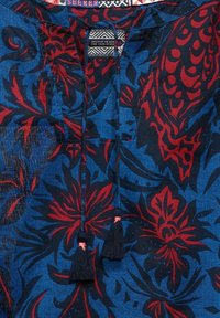 Tunique à motifs floraux bleus et rouges avec un col en V, dotée d'une fermeture à nouer et de deux pompons foncés. Fabriquée en tissu léger.