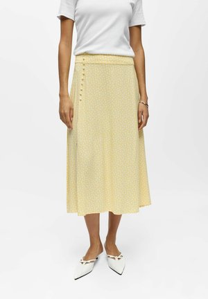 Femme portant un haut blanc à manches courtes, une jupe midi jaune à pois blancs avec des boutons sur le côté, et des chaussures blanches à bout pointu.