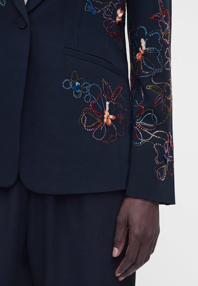 Μανίκι και τσέπη μπλέιζερ ναυτικού διακοσμημένα με πολύχρωμη κεντητή λουλουδένια διακόσμηση με χάντρες, εμφανίζονται με αντίστοιχο μπλε παντελόνι και χέρι με σκούρο δέρμα.